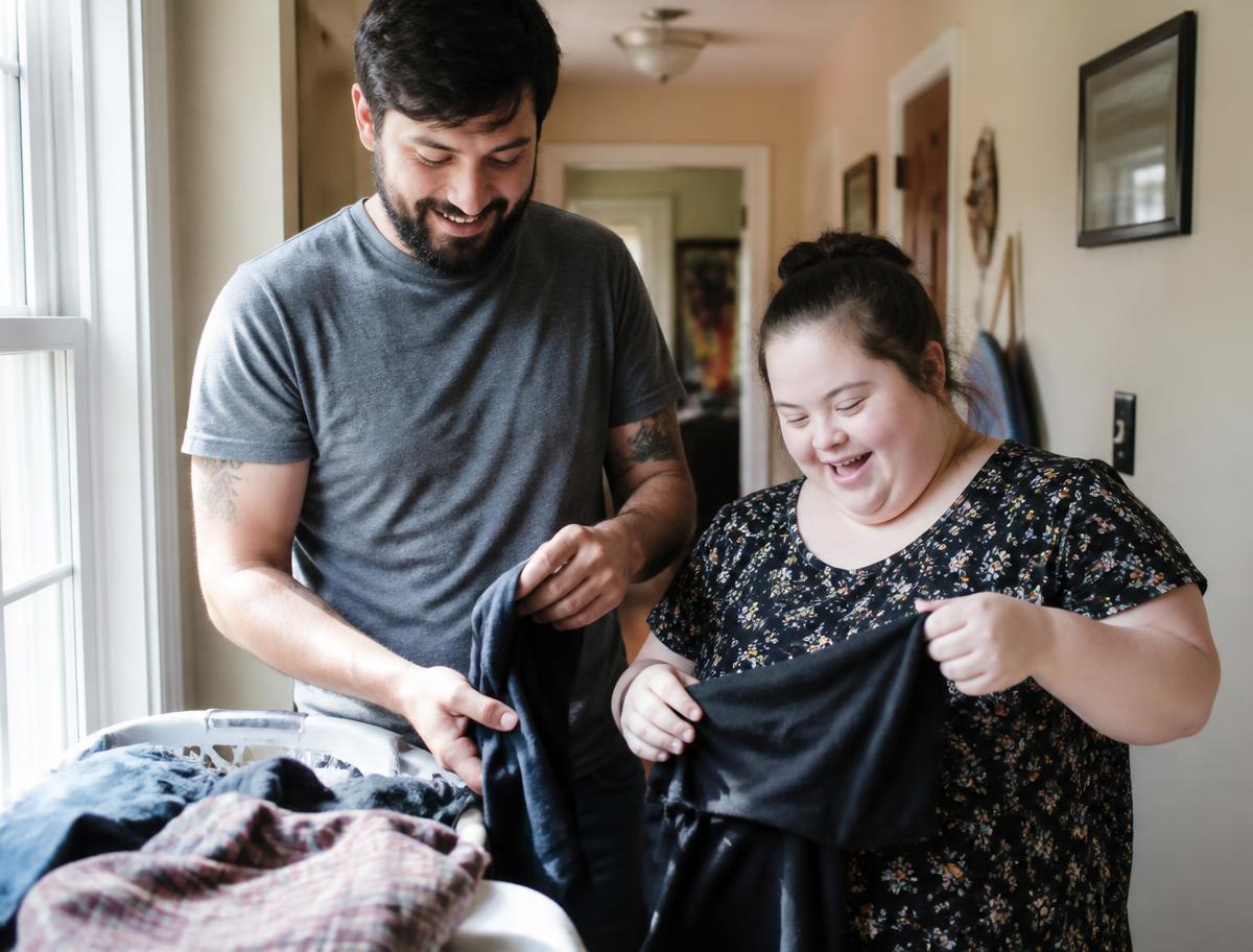 An adult brother and sister folding laundry together in a sunlit hallway at home
