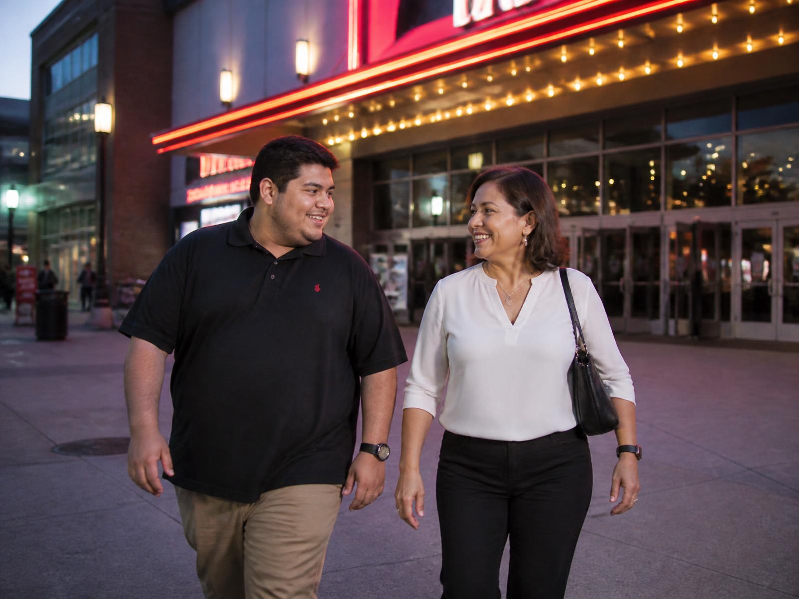 A young Hispanic man and his aunt walking side by side into a movie theater at a Maryland mall in the evening