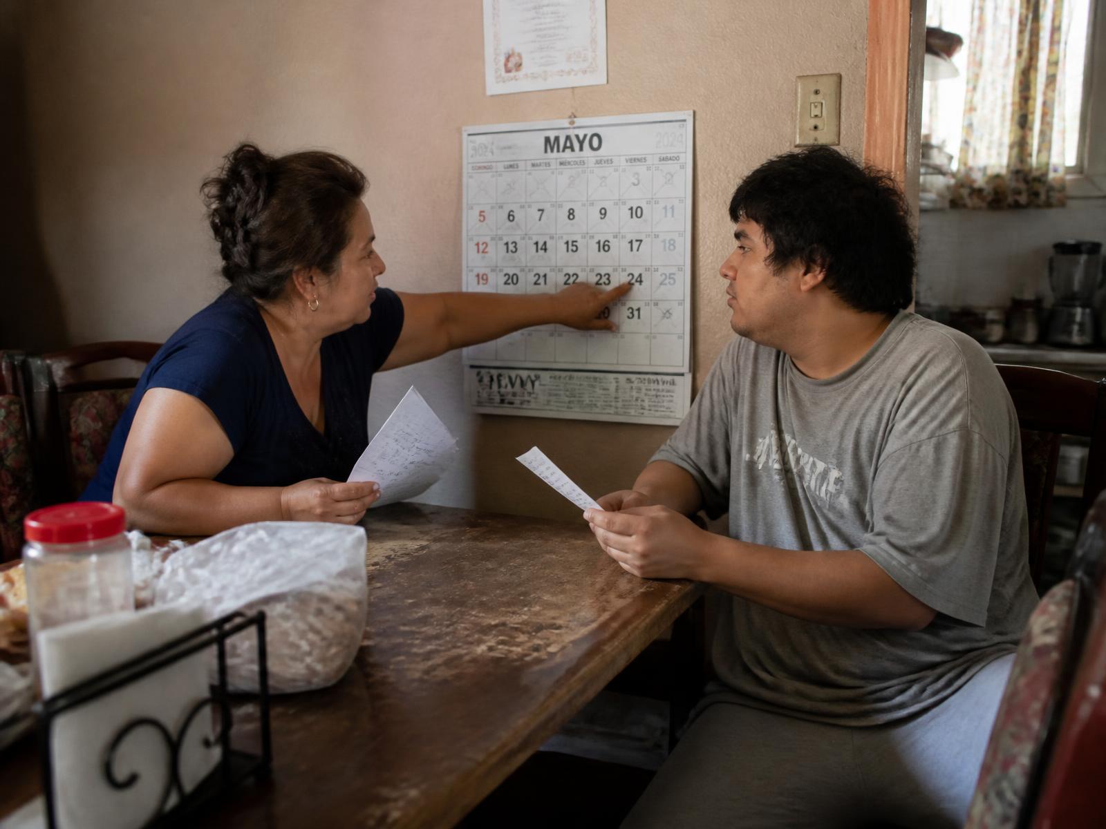 A Hispanic mother and her adult son reviewing a wall calendar and handwritten notes at their kitchen table